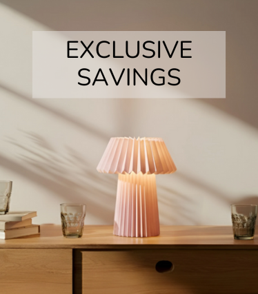 Bledě růžová stolní lampa na dřevěném příborníku, obklopená sklenicemi a knihami v teplém slunečním světle.