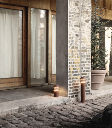 Venkovní scéna se dvěma moderními válcovými světly umístěnými vedle kamenného chodníku a zděno-betonového sloupu. Za nimi jsou skleněné dveře s dřevěným rámem, lehké závěsy a velká stálezelená rostlina v květináči. Teplá záře světel vytváří útulnou a příjemnou atmosféru.